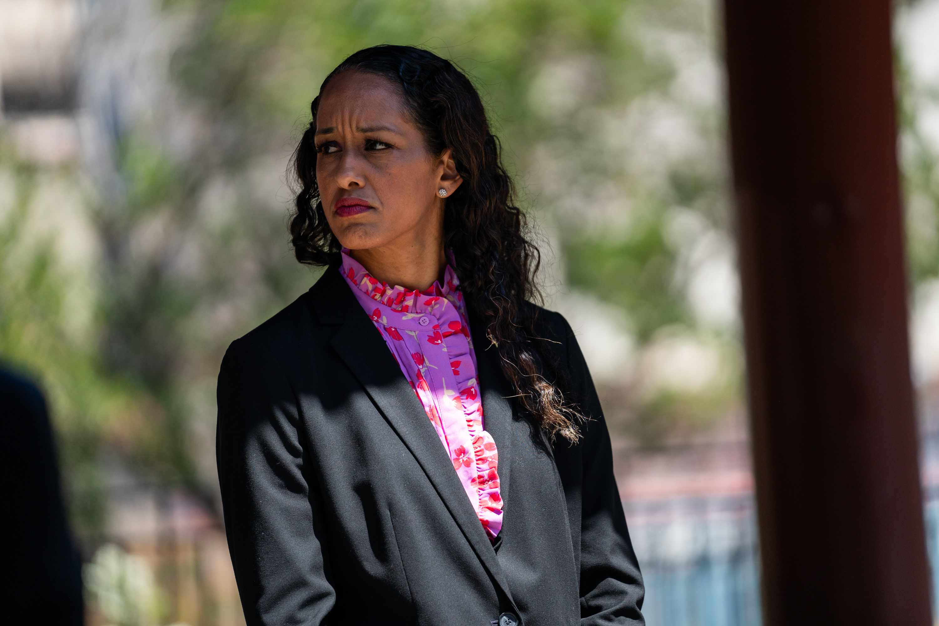 A person wearing a black blazer and a pink floral blouse is standing outdoors with blurred greenery in the background, looking serious and to the side.