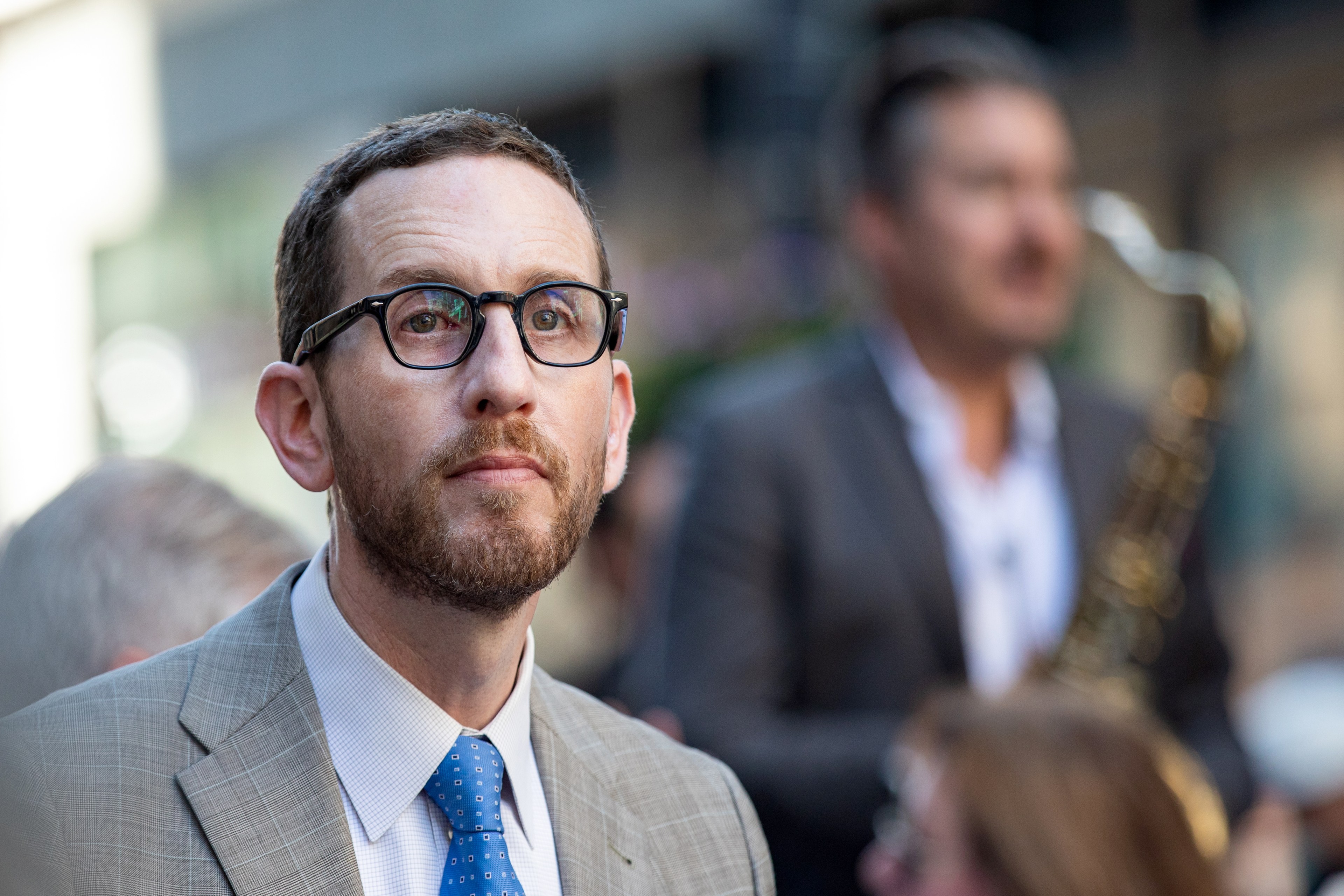 A man with glasses and a beard looking thoughtful, wearing a suit and tie, blurred saxophonist in the background.