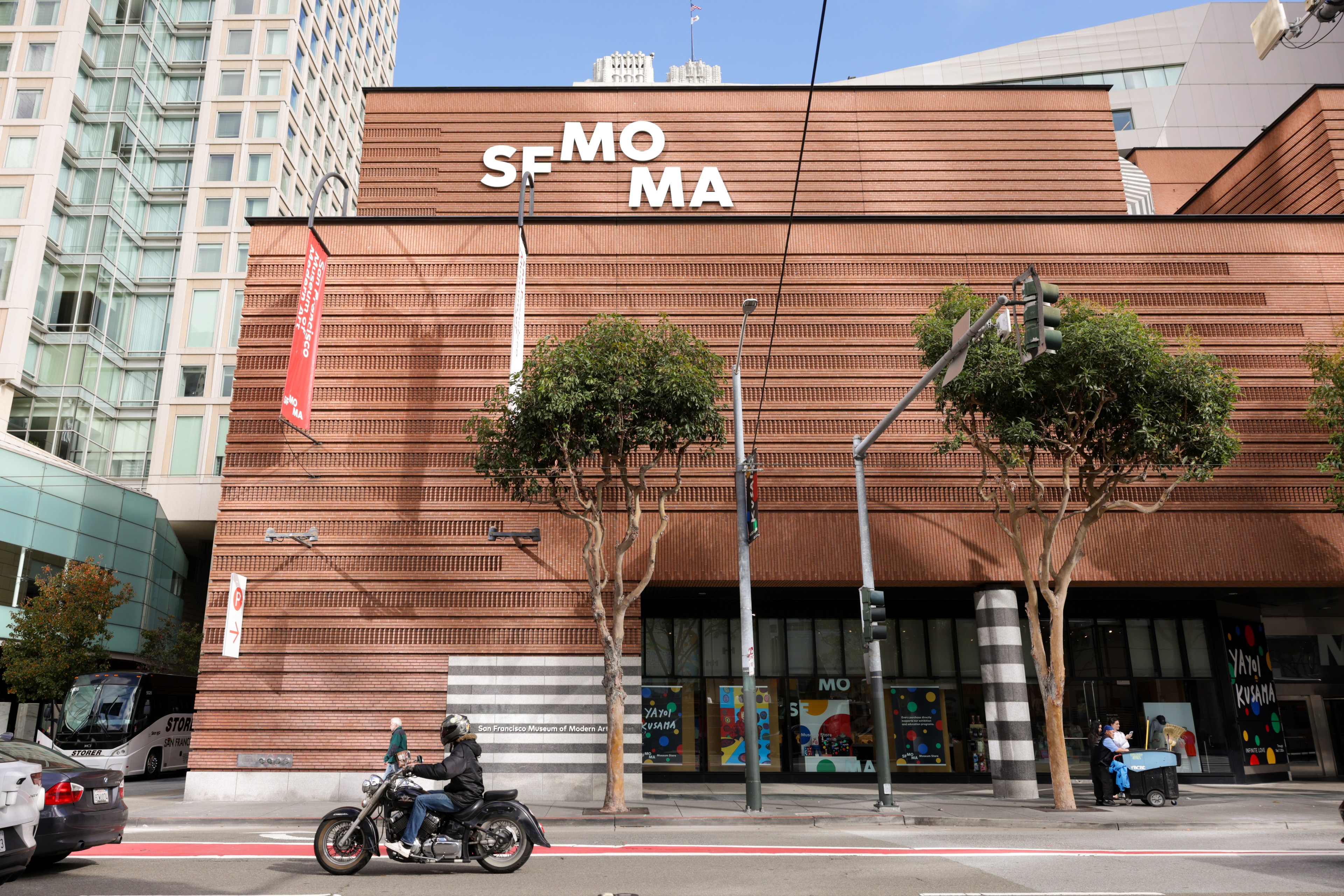 A museum stands behind trees and a motorcyle on the street.