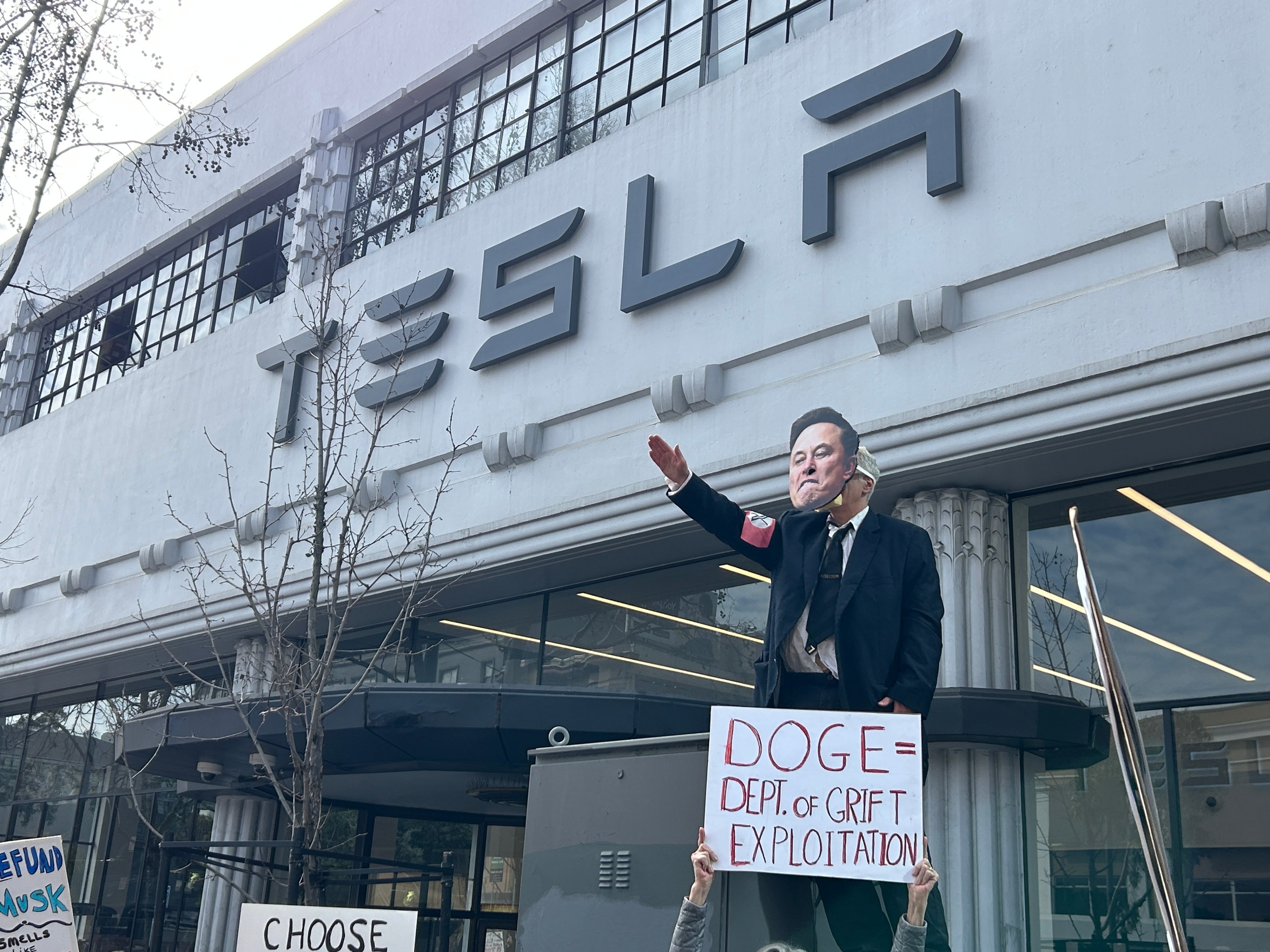 The image shows a Tesla storefront with a mannequin in a suit wearing a mask. People hold protest signs, with the largest reading "DOGE = DEPT. OF GRIFT EXPLOITATION."