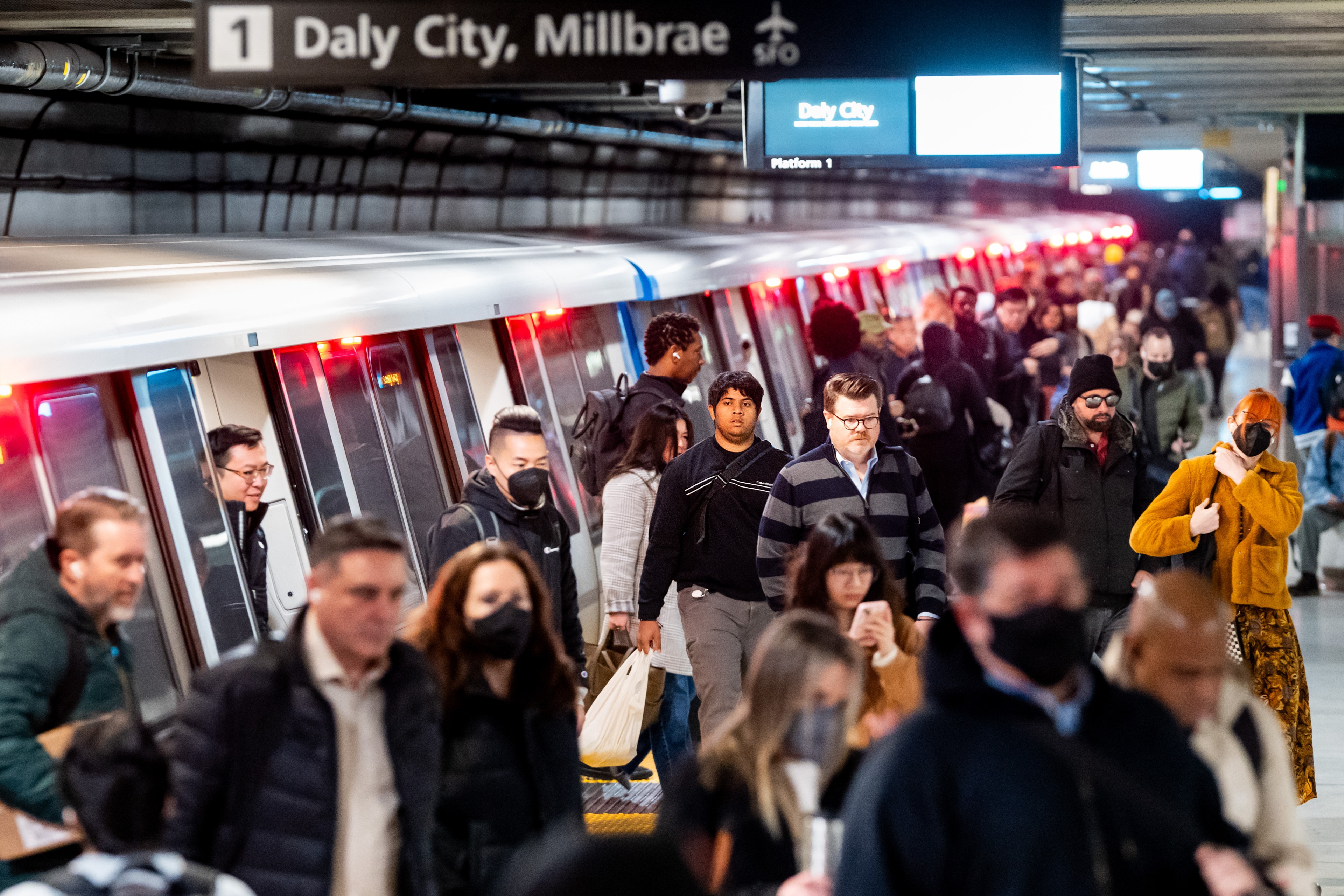 A busy subway platform filled with people, some wearing masks. A train is stopped with open doors. Signs above indicate destinations like Daly City and Millbrae.