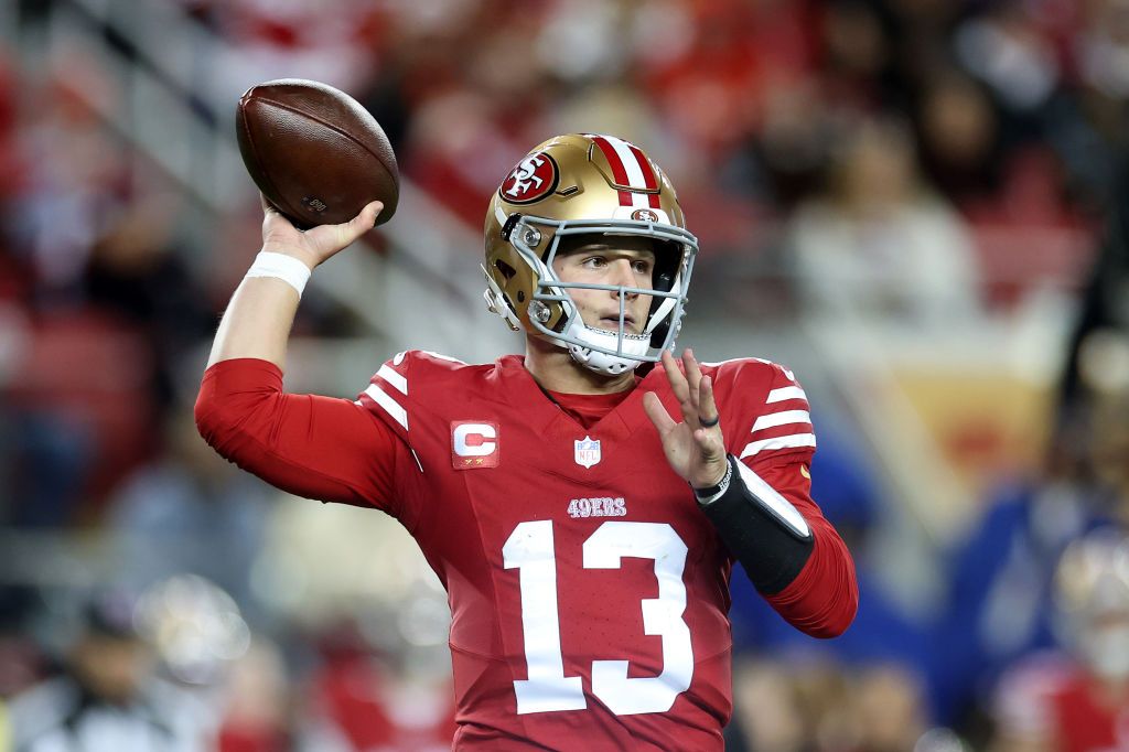 A football player in a red and gold uniform, with the number 13, is mid-throw holding a football in his right hand. He's wearing a helmet, and the background is a stadium.