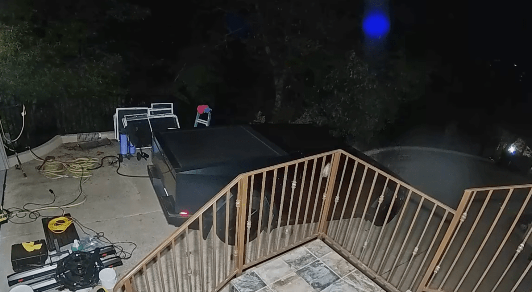 The image shows a nighttime scene of a cluttered patio with equipment, hoses, and stacked items near a railing. A parked trailer is visible in the background.