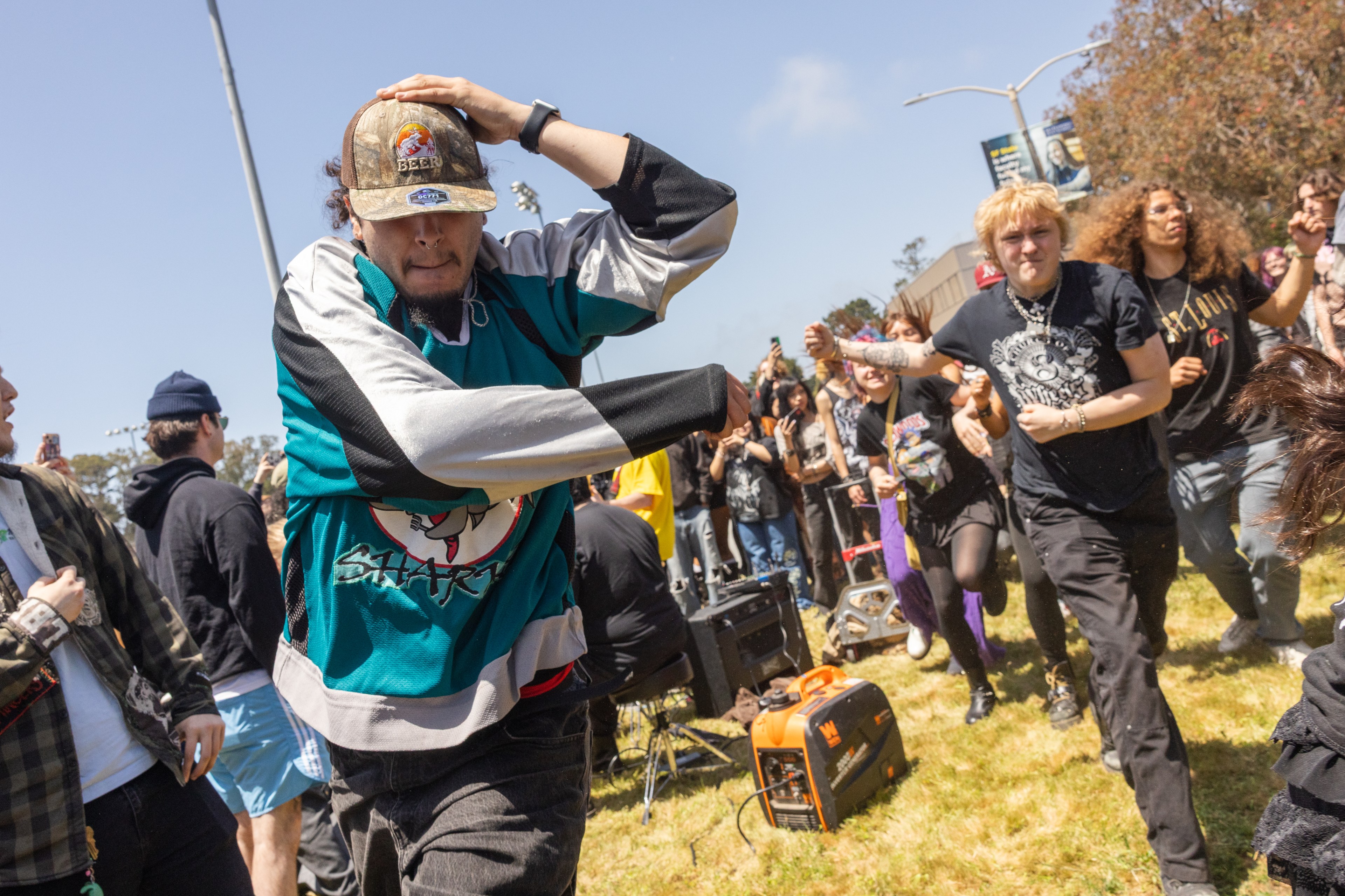 A lively outdoor scene shows a group of people dancing energetically. One person wears a teal jersey and cap, while others in dark clothing move actively in the background.