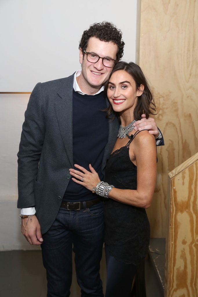 A man in glasses and a gray jacket stands with a smiling woman in a black dress and jewelry, posing closely together inside a room with wooden panels.
