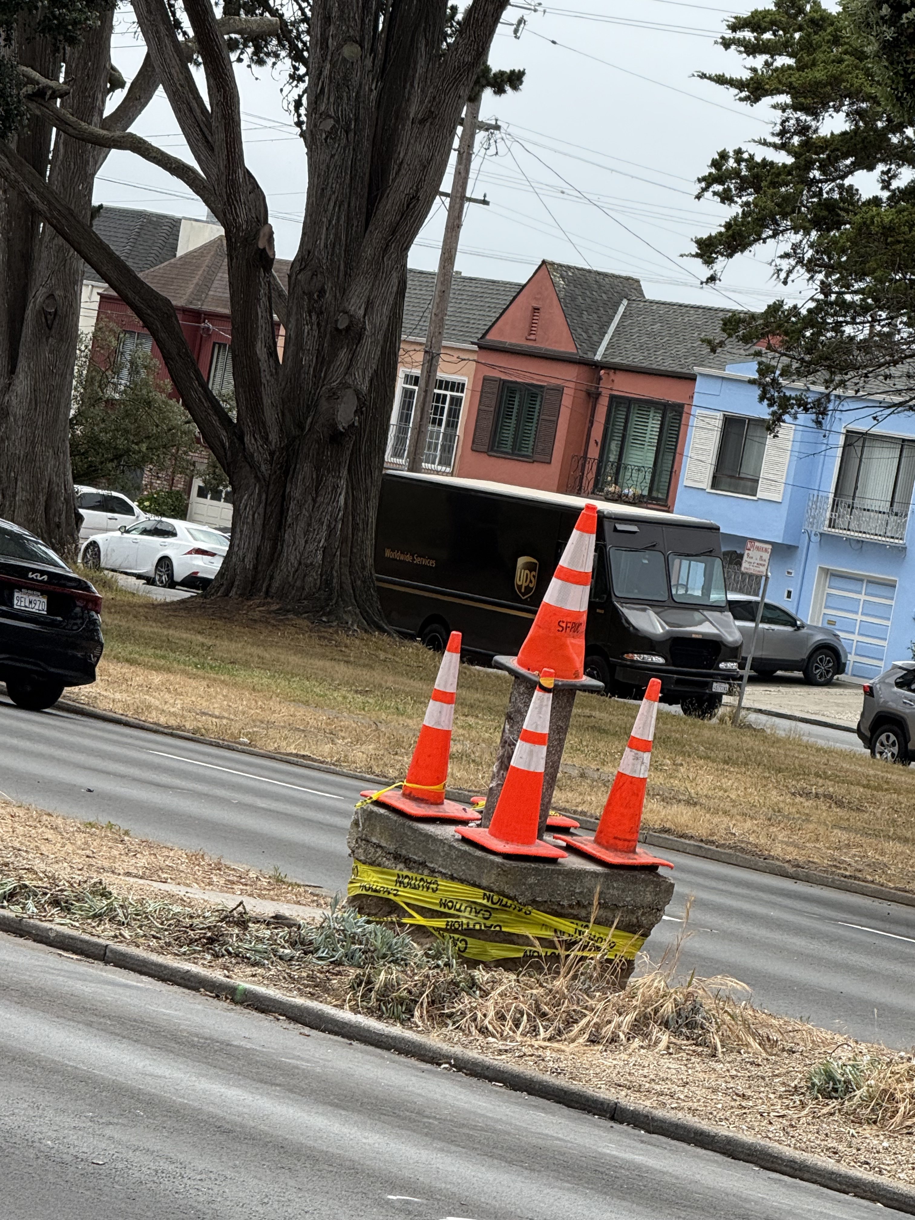 A concrete block with orange cones and caution tape is on a grassy area near a street. In the background, there's a UPS truck and colorful houses.