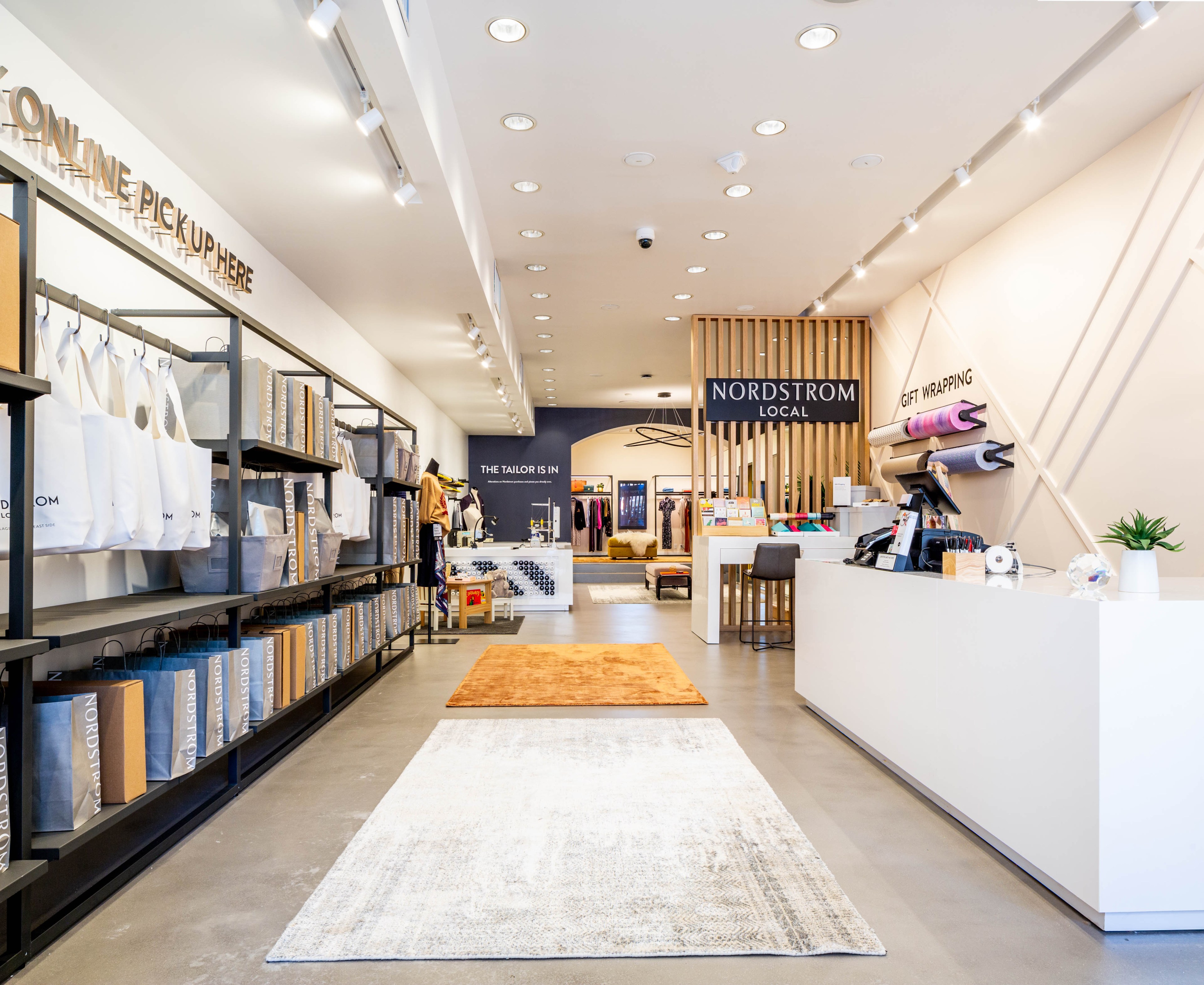 A sleek store interior with white walls features areas for online pickups, Nordstrom Local services, tailoring, and gift wrapping. Shelves display bags and boxes.