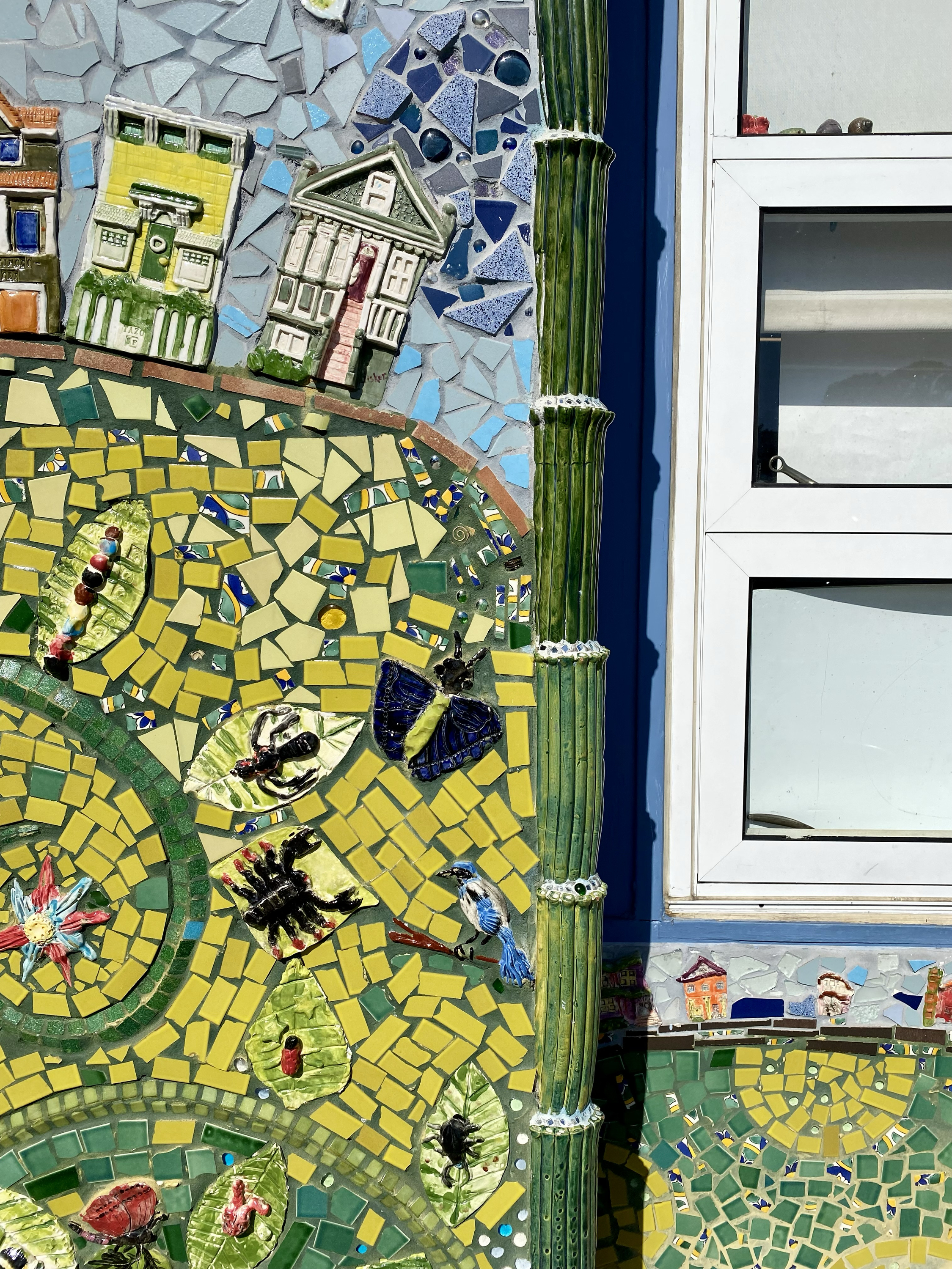The image shows a colorful mosaic featuring small buildings, insects like a butterfly and a scorpion, and a bird, with a tiled pattern of yellow, green, and blue.