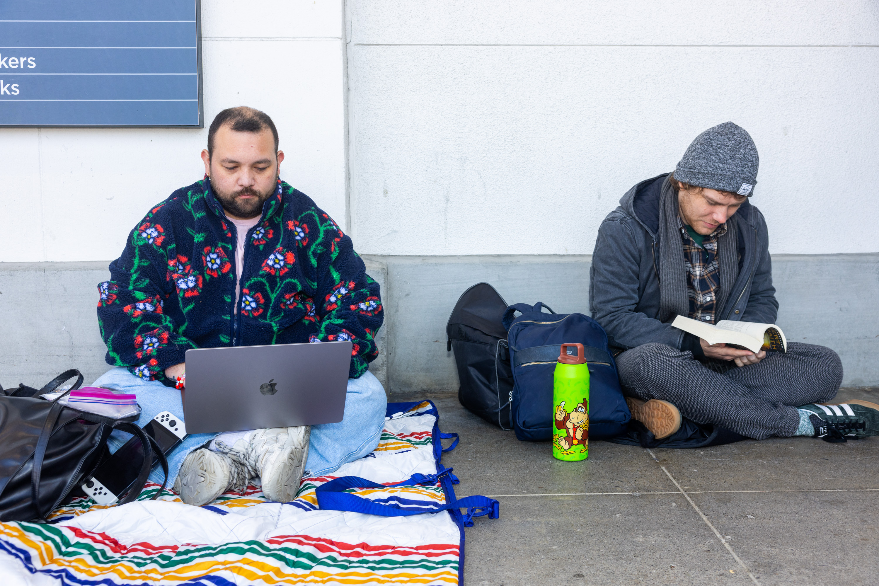Two people sit on the ground near a wall. One types on a laptop with colorful attire; the other reads a book, dressed warmly. A bright water bottle is nearby.