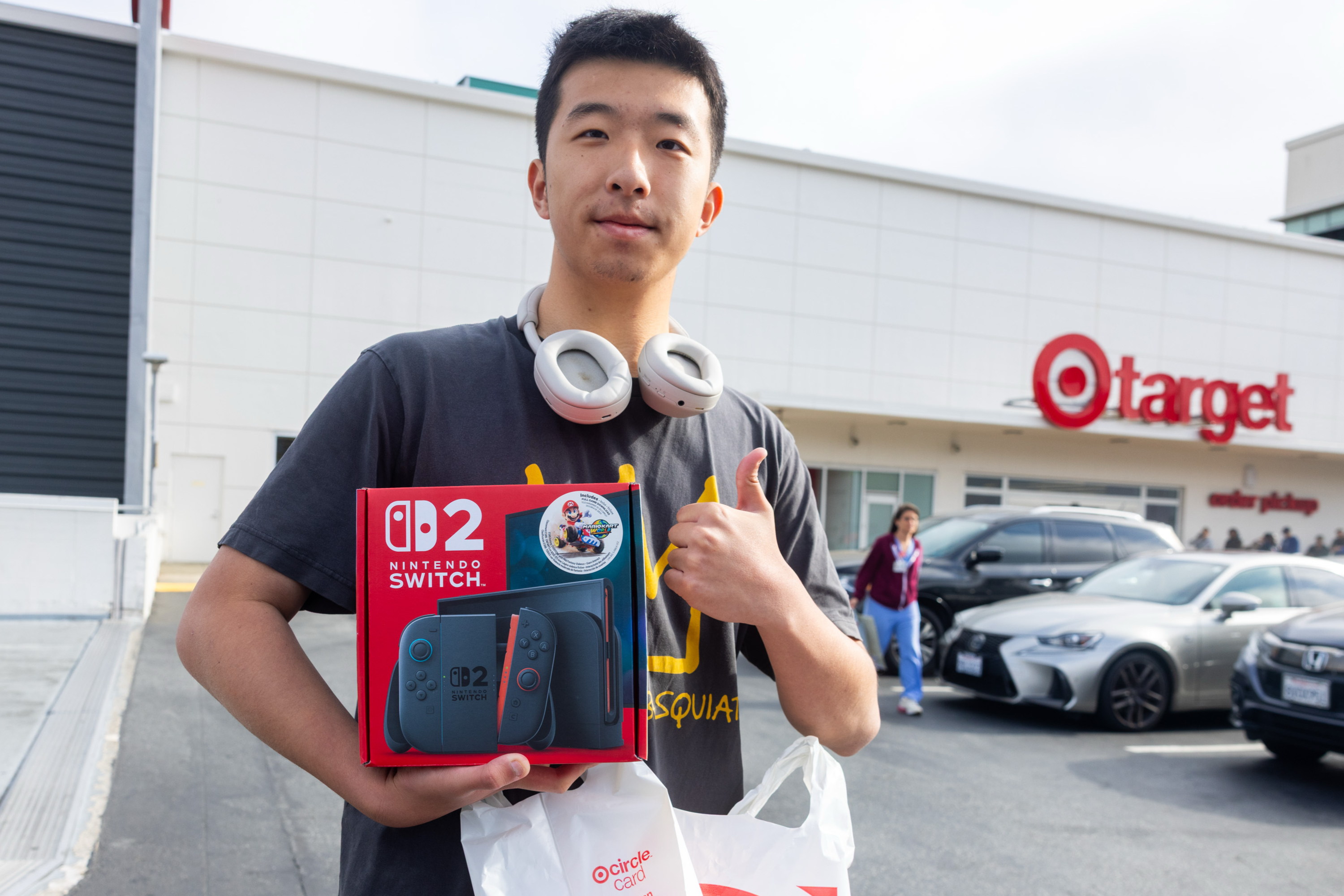 A person stands in a parking lot outside Target, holding a Nintendo Switch OLED box, wearing headphones and giving a thumbs-up. Cars and another person are in the background.