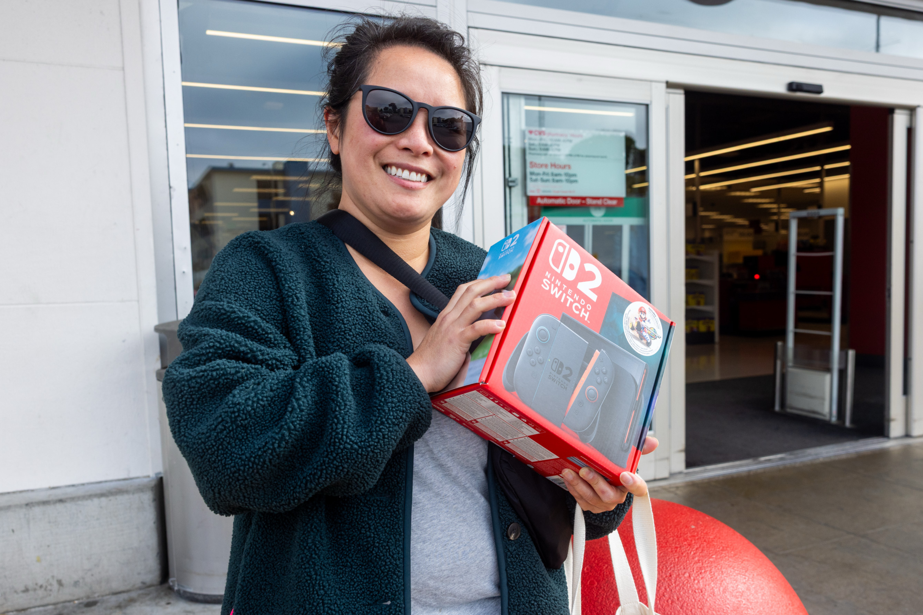 A person in sunglasses is smiling and holding a boxed Nintendo Switch OLED outside a store, near automatic doors.